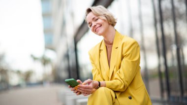 Gülümseyen sarışın kadın elinde cep telefonuyla şehirde oturuyor.