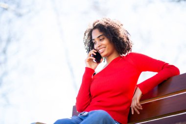 Kırmızı kazaklı, gülümseyen Afro-Amerikan kadın parkta bir bankta oturuyor, dışarıda cep telefonuyla konuşuyor..