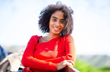 Afro-Amerikan bir kadının portresi. Bankta eğilmiş, kırmızı bir kazakla gülümsüyor..