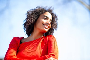 Kırmızı kazaklı Afro-Amerikan bir kadının yakından çekilmiş portresi. Dışarıda, güneş ışığında gülümsüyor..