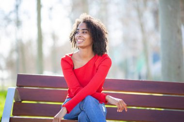 Afro-Amerikalı bir kadın bankta bağdaş kurup otururken gülümsüyor. Günlük kot pantolon ve kırmızı kazak giyiyor..