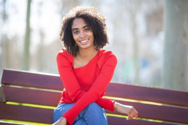 Gün ışığında bankta otururken kırmızı süveter giyen genç bir kadın gülümsüyor..