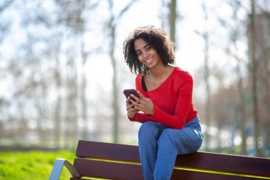 Kırmızı kazaklı, gülümseyen bir kadın açık hava bankında akıllı telefonuyla oturuyor..