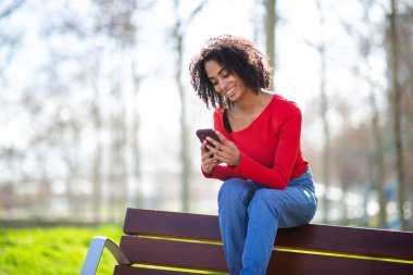 Parktaki ahşap bir bankta otururken telefonuna bakan genç bir kadın..