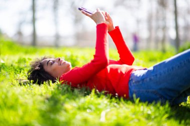 Genç siyahi kadın çimlerin üzerinde uzanıyor, gülümsüyor ve telefon ediyor, parkta boş vakitlerin tadını çıkarıyor..