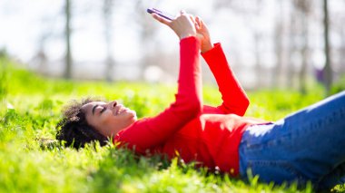 Gülümseyen genç siyahi kadın çimenlerin üzerinde uzanıyor, telefonu suratının üstünde tutuyor, dışarıda eğleniyor..