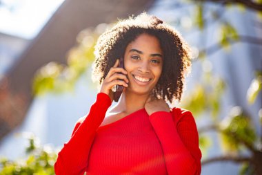 Açık havada akıllı telefondan konuşurken gülümseyen, kırmızı kazaklı, kendinden emin genç bir kadın..