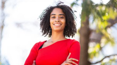 Kırmızı kazaklı, kolları çaprazlanmış, dışarıda duran gülümseyen genç bir kadın..