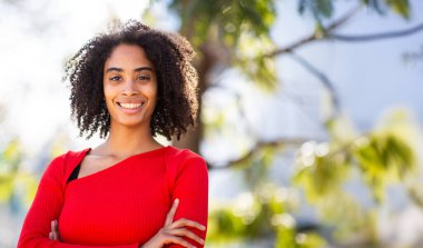 Kollarını kavuşturup gülümseyen genç bir kadın. Açık havada kendine güvenen bir duruş..