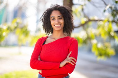 Gülümseyen kırmızı kazaklı genç kadın dışarıda poz veriyor, sakin bir ifade ve kolları bağlı..