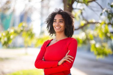 Kırmızı kazaklı, gülümseyen, kolları bağlı, açık havada duran siyah bir kadın.