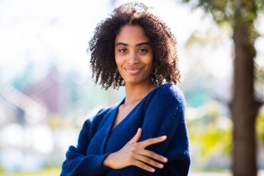 Kollarını kavuşturup gülümseyen, koyu mavi bir kazak giyen, açık havada duran genç bir kadın..