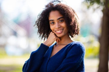 Açık havada mavi süveterli bir kadın, bir eli çenesinde hafifçe gülümsüyor.