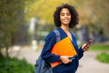 Gülümseyen Afrikalı Amerikalı kadın dışarıda telefon ve klasör tutuyor.