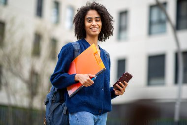 Telefon ve klasörle gülümseyen kendine güvenen siyah kız öğrenci.