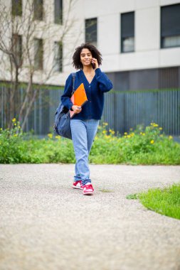 Telefon ve klasör taşıyan öğrenci güvenli bir şekilde dışarıda yürüyor.