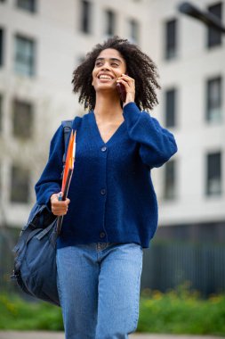 Gülümseyen Afrikalı Amerikalı öğrenci dışarıda telefonla konuşuyor.