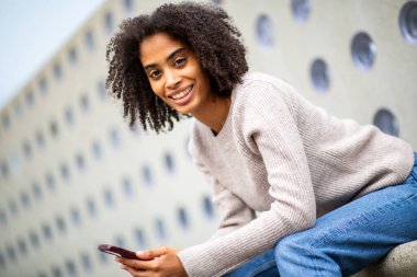 Dışarıda akıllı telefonuyla oturan gülümseyen genç bir kadın.