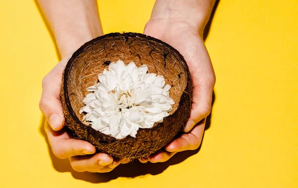 Female hands hold a flower in a coconut on a yellow background. Place for text flat lay. Spa salon, beauty treatments. Place for text