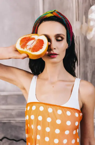 Profesyonel makyajlı güzel esmer kız bir gözünü greyfurtla kapatıyor. Çevre dostu cilt bakım ürünlerinin reklam afişi. Sabah güzellik rutini. Kozmetik bakım. vitamin