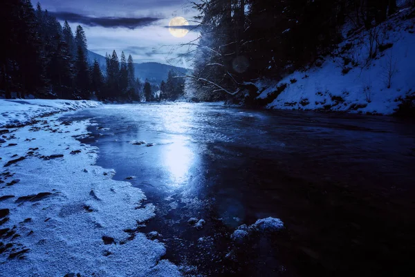 tereblya river in winter in full moon light. snow covered shore with coniferous forest on the shore. mountains beneath a cloudy sky on a frosty night in the distance. carpathian Christmas vacations in white season