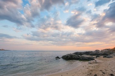 Şafak vakti kumlu ve taşlı sıcacık deniz kıyısında. Yoğun bulutlarla dolu bir gökyüzünün altında ufukta güzel bir manzara var. Yaz tatili sezonu
