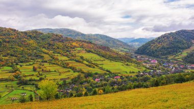Sonbaharda Karpat dağlarının kırsal kesiminde. Uzak vadideki köy. Tepelerdeki ağaçlar sonbahar renklerinde. Bulutlu bir gökyüzünün altındaki uzak dağ sıraları