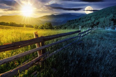 Alacakaranlıkta güneş ve ay olan kırsal alan. Çit çayırın yanında, ağaçlar da yamaçta. Gündüz ve gece zaman kavramını değiştirir. Gizemli kırsal manzara Sabah ışığında