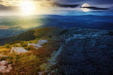 Alacakaranlıkta güneş ve ay olan dağ manzarası. Yamaçta çimenlerde taşlar var. Gündüz ve gece zaman kavramını değiştirir. Gizemli kırsal manzara Sabah ışığında