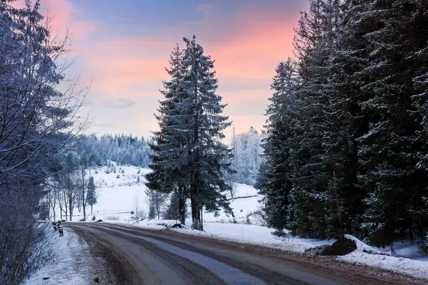 Kışın gün batımında asfalt yol orman kırsalından geçiyor. Karda ağaçlar ve tepeler