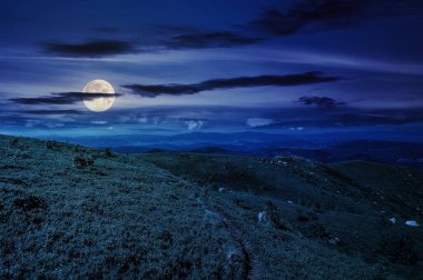 Geceleri yaz manzarası. Bulutlu gökyüzünün altındaki dağ sırtındaki yamaçtaki çayır boyunca yol. Dolunay ışığında harika bir kır manzarası