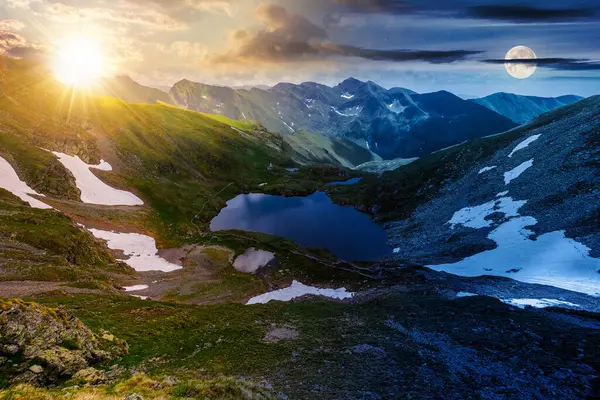 summer solstice in mountains of romania. beautiful landscape with capra lake and snow on the hills beneath a sky with sun and moon. day and night time change concept