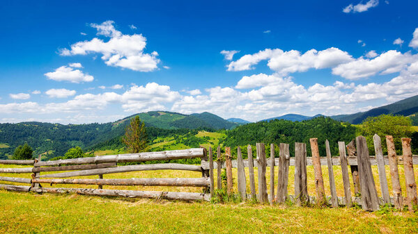 wooden fence on the meadow. mountainous rural landscape of transcarpathia, ukraine in summer. carpathian countryside with forested rolling hill beneath a blue sky with white fluffy clouds on a sunny day at high noon
