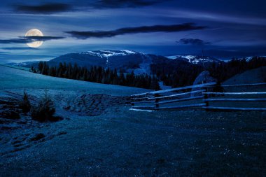 Geceleri kırsal alanda çitlerle çevrili. Dolunay ışığında ormanlık tepeleri olan dağlık bir kır manzarası.