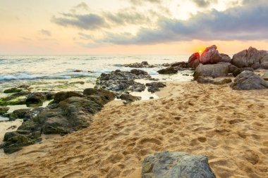 Yaz tatili manzarası. Gün doğumunda dramatik deniz manzarası. Sakin dalgalar sahili kum ve kayalarla yıkıyor. Ufukta güneş ve bulut olan güzel gökyüzü manzarası