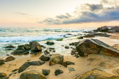 Yaz tatili manzarası. Gün doğumunda dramatik deniz manzarası. Sakin dalgalar sahili kum ve kayalarla yıkıyor. Ufukta güneş ve bulut olan güzel gökyüzü manzarası
