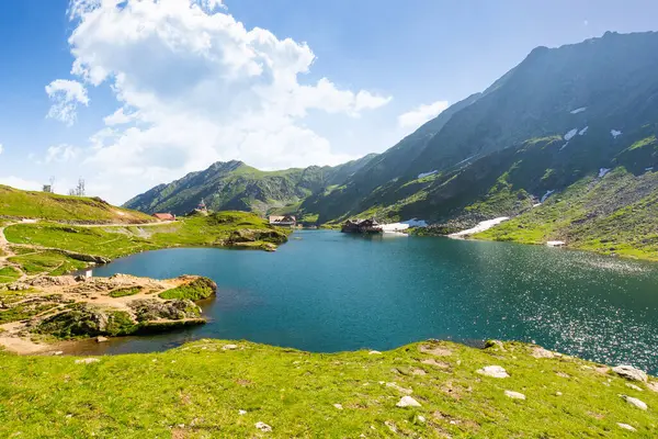 Romanya 'nın Balea Gölü. İbnelerin göletli dağlarının alp manzarası. Karpatların üstündeki mavi gökyüzünde bulutlarla güneşli bir hava. Yazın yamaçta kar ve çimenler. Cennet gibi bir yer.