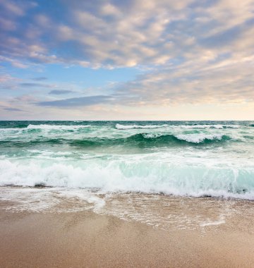 Sabahın köründe deniz manzarası. Kötü tatil sezonu geçmişi. Suyun doğal gücü. Yazın bulutlu gökyüzü ile fırtınalı bir hava. Deniz dalgası kıyıya çarpıyor. ıslak kum