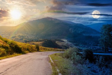 Alacakaranlıkta güneş ve ay ile dağ geçidi. Tepeden aşağı inen yol. Bulutlu gökyüzünün altında tepenin önündeki vadi manzarası. Gündüz ve gece zaman kavramını değiştirir