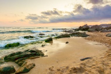 Yaz tatili manzarası. Gün doğumunda dramatik deniz manzarası. Sakin dalgalar sahili kum ve kayalarla yıkıyor. Ufukta güneş ve bulut olan güzel gökyüzü manzarası