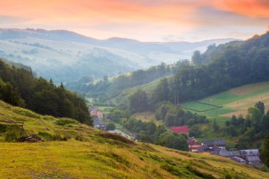 Şafak vakti gökyüzü olan kırsal vadi manzarası. Yazın sabah doğası. Dağdaki yeşil çayır ve tepedeki orman. Manzaralı seyahat manzarası. Açık hava turizmi için çimenleri olan güzel bir kırsal alan.