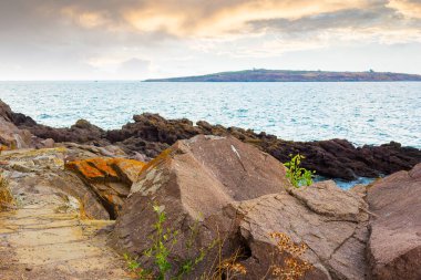 Sahildeki deniz taşları. Sahilde taşlarla birlikte doğayı gezmek. Ufuktaki ada. Deniz kenarında güzel bir yaz tatili. Mavi suyun sakin yüzeyi ve deniz manzaralı akşam gökyüzü