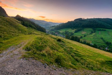 Yazın kırsal alan. Karpatya menzili. Şafakta vadideki köy. Kızıl gökyüzünün altındaki tepede orman ve yeşil çayır. Ukrayna dağlarının manzaralı manzarası.