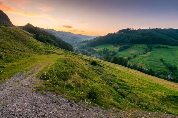Yazın kırsal alan. Karpatya menzili. Şafakta vadideki köy. Kızıl gökyüzünün altındaki tepede orman ve yeşil çayır. Ukrayna dağlarının manzaralı manzarası.