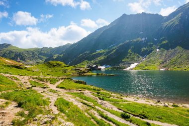Romanya 'nın Balea Gölü. İbnelerin göletli dağlarının alp manzarası. Karpatların üstündeki mavi gökyüzünde bulutlarla güneşli bir hava. Yazın yamaçta kar ve çimenler. sakin su yüzeyi