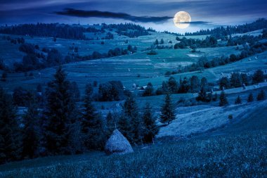 Geceleri kırsal dağ manzarası. Köyün yakınındaki yeşil tepede saman yığını. Alp manzaralı Fantezi sırtında dolunay ışığında