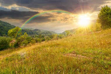 Yazın gün batımında bulutlu gökyüzü ile kırsal alan. Çimenli tepedeki orman. Gökkuşağının altında akşam ışığında bulutlu hava tahmini. Dağlardaki uzak kırsal manzara