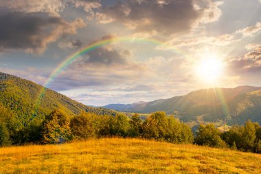 Sonbaharda günbatımında tepenin üzerinde ormanla kaplı dağ manzarası. Akşam ışığında kırsal vadiye güzel bir manzara. Gökkuşağı ve gökyüzü altında bulutlu dağlık arazi manzarası