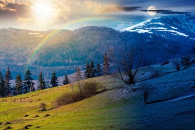 Bahar ekinoksunda dağlık kırsal alan. Gündüz ve gece değişiyor. Bir tepenin yamacındaki çimen tarlası. Uzakta karlı tepeli dağ sırtı Alacakaranlıkta güneş ve ay ile