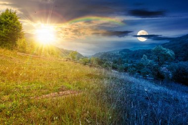 Yazın bulutlu gökyüzü olan kırsal alan. Gündüz ve gece zaman kavramını değiştirir. Alacakaranlıkta güneş ve ay olan çimenli tepede bir orman. Bulutlu hava tahmini. Dağdaki uzak kırsal manzara